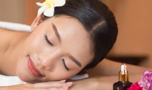 A guest resting peacefully on a white towel with a frangipani in her hair, a bottle of Sapaiya oil beside her